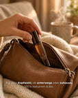 Hand zieht die NAYURE Head Relief Roll-on Flasche aus einer braunen Ledertasche, im Hintergrund eine Decke, eine dampfende Tasse und eine Kerze