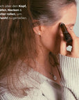 Frau rollt den NAYURE Head Relief Roll-on über die Schläfe, im Profil mit langen braunen Haaren und beigem Strickpullover